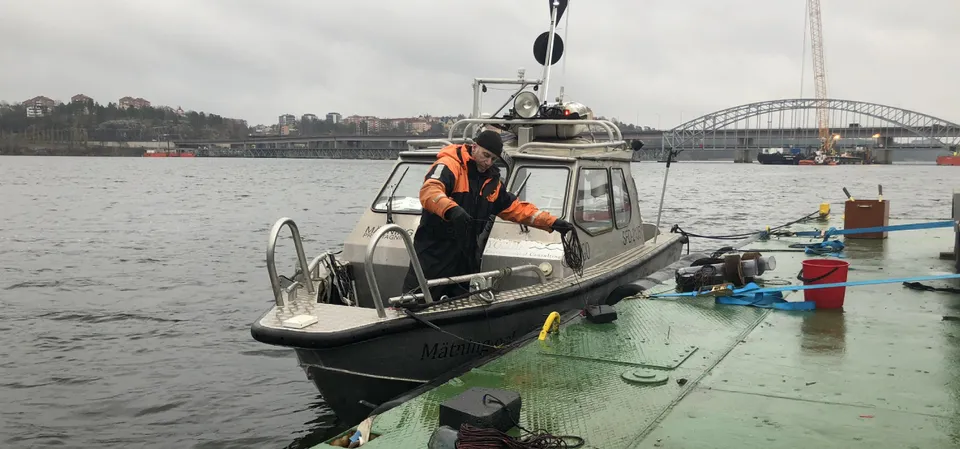Yoldias mätbåt förtöjd vid en ponton. En person i flytoverall arbetar på fördäck och en bågbro syns i bakgrunden.